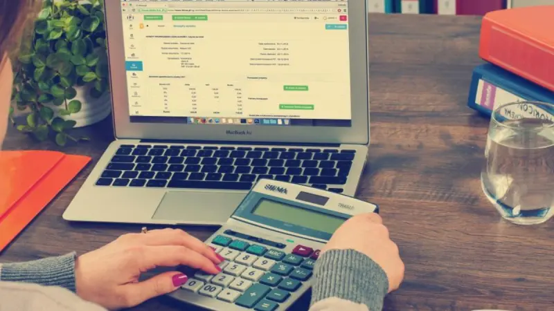 A person working a desk calculator beside a laptop showing an invoice